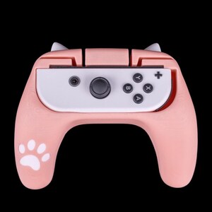 Peut inclure: Une manette Nintendo Switch rose et blanche avec des oreilles de chat et une empreinte de patte sur le côté. La manette est conçue pour être utilisée avec une console Nintendo Switch.