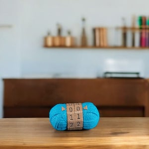 Könnte beinhalten: Ein knallblauer Wollknäuel mit einem braunen Etikett, auf dem "0 0 1 1 2 2" steht. Der Wollknäuel liegt auf einer Holzoberfläche.