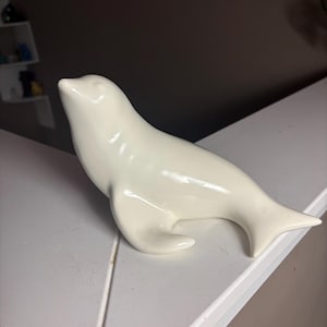 以下が含まれることがあります： 休息姿勢をとるオフホワイトの陶器製アザラシの置物。頭を上に向けており、流線型の体と横に伸びた尾びれが特徴で、全体に光沢のある滑らかな仕上げが施されています。表面は反射する質感で、シンプルで洗練されたデザインの置物です。