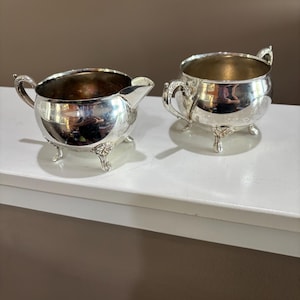 May include: A silver-toned creamer and sugar bowl set featuring polished metallic surfaces, curved handles, and ornate tripod feet. Both vessels have rounded bodies and wide openings, reflecting light across their smooth, reflective exterior finishes.