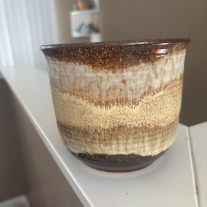 Peut inclure: Une tasse en céramique cylindrique présentant un émail aux tons multiples avec des bandes horizontales marron foncé, crème et beige. La surface affiche un aspect texturé et superposé avec de légères coulures verticales et un bord arrondi.
