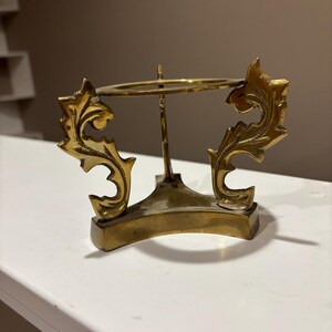 May include: A brass-coloured decorative stand with an ornate, openwork design. The stand features a circular top supported by a central post and a base with two decorative, leaf-like elements. The stand is designed to hold a small object.