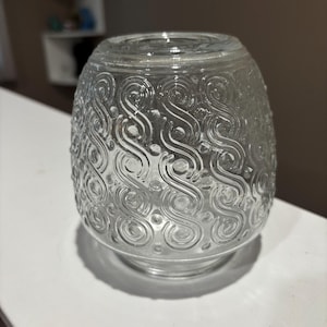 May include: Clear glass light fixture shade with an egg-like shape. The shade features a repeating pattern of swirling, embossed designs. The glass is transparent, allowing light to pass through. The shade sits on a white surface.