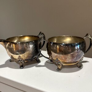 May include: A silver-plated creamer and sugar bowl set. The creamer has a spout and the sugar bowl has two handles. Both pieces have ornate feet and a tarnished finish, suggesting age and use. The interiors have a golden hue.