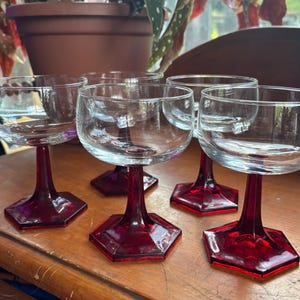 Könnte beinhalten: Set mit sechs Vintage-Champagner-Coupe-Gläsern. Jedes Glas hat eine klare Schale auf einem rubinroten Stiel und einem sechseckigen Fuß. Die Gläser sind auf einer Holzoberfläche angeordnet und zeigen ihr elegantes Design.