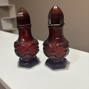 Può includere: Due saliere e pepiere abbinate in vetro rosso rubino. Ogni shaker ha un design decorativo e strutturato e una parte superiore a punta. Gli shaker sono presentati su una superficie bianca.