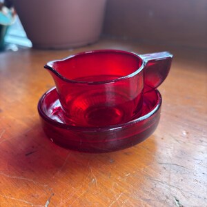 以下が含まれることがあります： 小さなルビーレッドのガラス製クリーマーとソーサーのセット。クリーマーには注ぎやすいようにハンドルと注ぎ口が付いています。ソーサーは丸く、クリーマーをしっかりと保持するためにわずかにくぼんでいます。セットは透明なガラス製です。