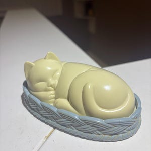 Peut inclure: Une figurine couleur crème représentant un chat endormi recroquevillé dans un panier ovale bleu clair à la texture tressée. Le chat a les yeux fermés et les pattes repliées près du visage, reposant dans une base bleue moulée imitant l'osier.