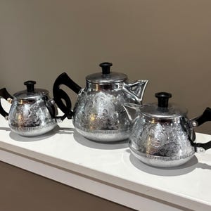 May include: Three silver teapots with black handles and lids. The teapots have an ornate, engraved floral design. The teapots are of varying sizes, with the largest in the center and the smallest on the left.