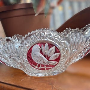 May include: A clear, cut-glass bowl with a red centre featuring a white bird and leaf design. The bowl has a scalloped edge and decorative handles. A decorative piece for serving or display.
