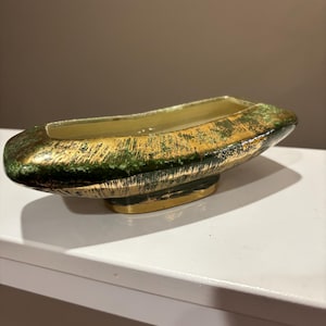 Op de afbeelding: Een decoratieve, langwerpige keramische schaal met een gouden binnen- en buitenkant. De buitenkant heeft een getextureerde, verweerde afwerking met groene en gouden accenten. De schaal staat op een kleine, goudkleurige basis.