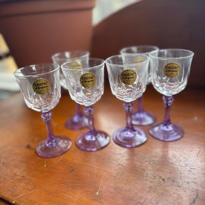 May include: Six clear crystal cordial glasses with purple stems are arranged on a wooden surface. Each glass has a gold label that reads "Cristal d'Arques". The glasses are in a row, with the stems and bases visible.