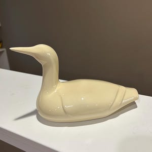 May include: A cream-colored ceramic duck figurine. The duck has a long neck and a pointed beak. The figurine is smooth and glossy, with subtle details. It is a decorative item.