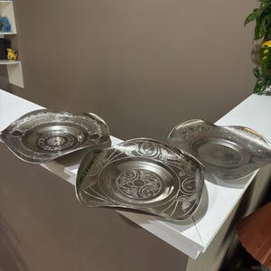 May include: Three silver-toned metal plates with wavy, irregular edges. Each plate features a central circular area surrounded by an intricate, etched floral and scrollwork pattern that extends towards the scalloped, raised rims of the dishes.