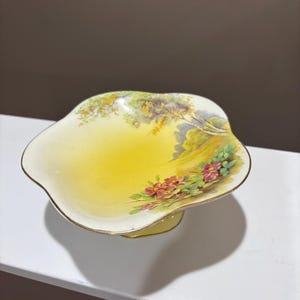 May include: A yellow and white footed dish with a scalloped edge. The dish features a landscape design with pink flowers, green foliage, and a gold rim. The dish is likely made of porcelain or ceramic.