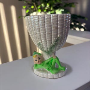 May include: A ceramic egg cup featuring a basket-weave design in off-white, with a green and white figure of a character in green attire reclining at the base. The figure has a green hat and is resting on a white base.