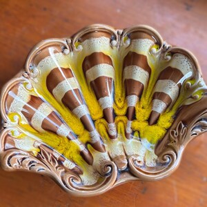 Peut inclure: Un plat en céramique en forme de coquillage, marron, jaune et blanc. Le plat présente un bord festonné et un motif de segments rayés bruns et blancs rayonnant du centre. L'intérieur est jaune.