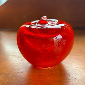 May include: A vibrant red glass apple figurine with a glossy finish. The apple has a stem and a smooth, rounded shape. The figurine is sitting on a wooden surface.