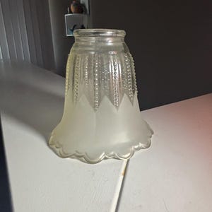 May include: A clear glass lamp shade with a bell shape, featuring a frosted finish on the lower half and a scalloped edge. The upper section displays vertical rows of raised beaded texture, transitioning into a smooth, frosted triangular pattern.