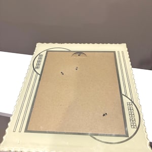 May include: Cream-colored picture frame with a scalloped edge and a rectangular opening. The frame has a decorative design in the corners and along the sides, with a light brown interior. The frame is made of a sturdy material.