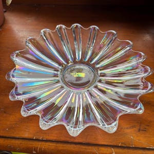 May include: A clear glass dish with a scalloped, flower-like edge. The dish has a central, slightly recessed area and displays an iridescent sheen, reflecting a spectrum of colours. It sits on a wooden surface.