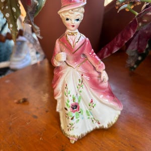 May include: A vintage porcelain figurine of a woman in a pink and white dress and hat. The figurine has gold accents and floral details on the dress. The woman is wearing a pink coat and a bow tie.