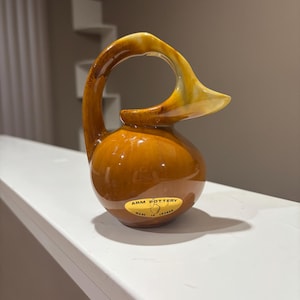 May include: A brown ceramic pitcher with a unique handle design. The handle curves upward and outward, transitioning from a light yellow to a darker brown. The pitcher has a rounded body and a label that reads "ARM POTTERY MADE IN CANADA."