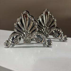 May include: Two silver-toned metal napkin holders featuring an ornate, symmetrical shell-like design with intricate scrollwork and floral accents at the base. The metallic finish is reflective, highlighting the raised relief patterns on each piece.