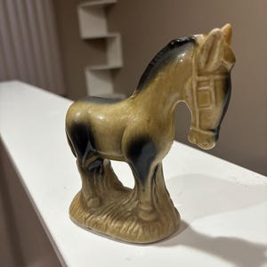 May include: A ceramic horse figurine in shades of tan, brown, and black. The horse has a detailed mane and harness, standing on a textured base. The figurine is a decorative item.