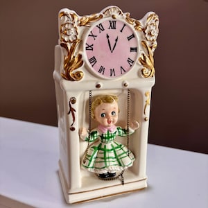 May include: A ceramic clock figurine featuring a cream-colored frame with gold accents and a pink clock face with black Roman numerals. A small figure with blonde hair wearing a green and white plaid dress hangs from chains inside the frame.