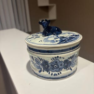 Może przedstawiać: Ceramiczne pudełko na drobiazgi z pokrywką, z niebiesko-białym kwiatowym wzorem. Pokrywka zwieńczona jest małą niebieską figurką. Pudełko ma cylindryczny kształt i jest ozdobione ręcznie malowanymi kwiatami.