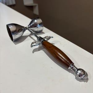 Könnte beinhalten: Ein doppelter Jigger aus Edelstahl mit einem dunkelbraunen Holzgriff. Der Jigger hat zwei gegenüberliegende Becher unterschiedlicher Größe, die durch einen Stiel verbunden sind. Der Griff hat dekorative Metallakzente.