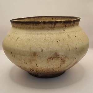 Op de afbeelding: Een grote, ronde, keramische pot met een lichtbeige glazuur en bruine vlekken. De pot heeft een brede, afgeronde basis en een licht uitlopende rand.