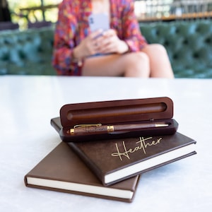 May include: A brown wooden pen with a gold trim and the name "Heather" engraved on it. The pen is in a brown wooden box with a brown leather notebook on top of it. The notebook has the name "Heather" written on it in gold lettering.