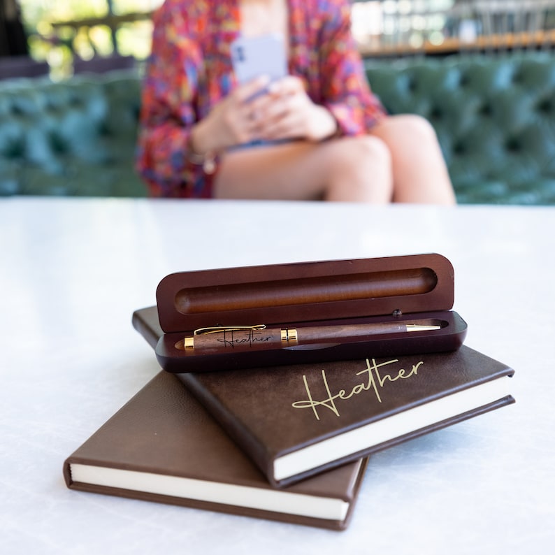 May include: A brown wooden pen with a gold trim and the name "Heather" engraved on it. The pen is in a brown wooden box with a brown leather notebook on top of it. The notebook has the name "Heather" written on it in gold lettering.