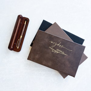May include: A stack of three brown leather-bound journals with gold lettering that reads "my dose" and a gold leaf design. A wooden pen case with a gold pen engraved with the name "Sophia" is in the upper left corner of the image.