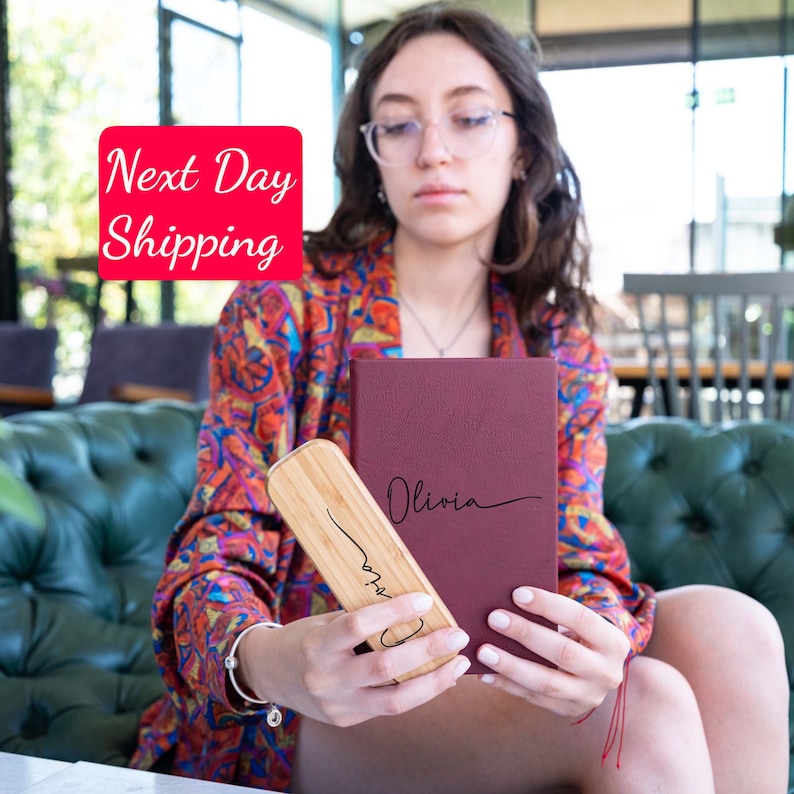 May include: A burgundy leather journal with the name "Olivia" embossed in gold lettering. The journal is being held by a person, along with a wooden pencil holder.