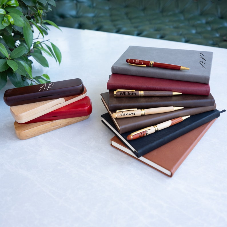 May include: A stack of six brown leather journals with gold-toned pens on top. Each pen has a different name engraved on it: Liam, William, James, Jack, and AP. The journals are stacked on a white surface.