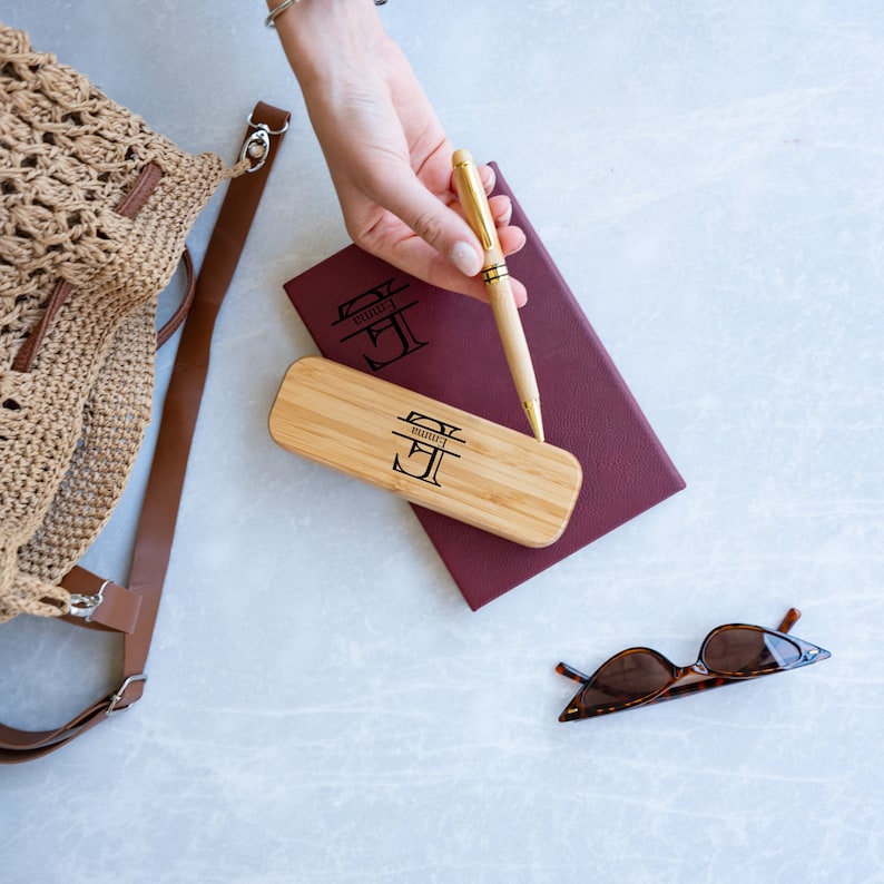 May include: A wooden pen case with a gold pen and the initials "E.M." engraved on the top. The pen case is sitting on a burgundy leather journal with the same initials engraved on the cover.