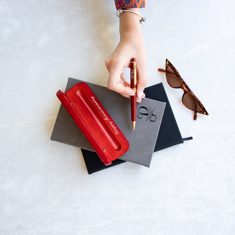 May include: A red wooden pen holder with the inscription "Happy anniversary" and a gold pen with the name "Peter" engraved on it. The pen is resting on a gray notebook with a black notebook underneath.
