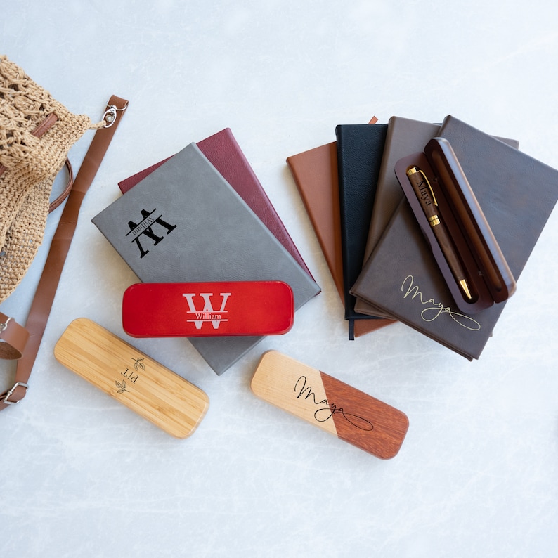 May include: A set of personalized stationery items, including a gray journal with the initials "YY" engraved, a red pen case with the name "William" and the letter "W" engraved, a wooden pen case with the name "Maya" engraved, and a brown leather journal with the name "Maya" engraved.