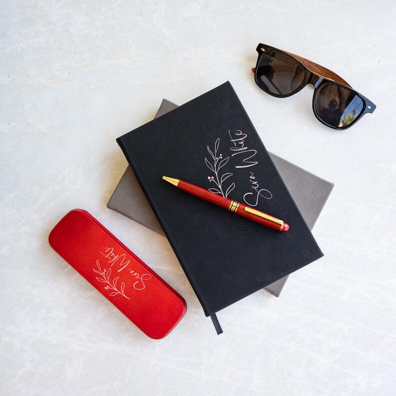 May include: A red pen case with a white floral design and the text "Save More" on a white background. A black notebook with a white floral design and the text "Save More" on a gray notebook. A pair of brown and black sunglasses.