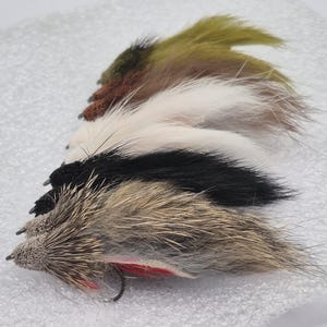 May include: A selection of fishing flies in shades of brown, black, white, and olive green. Each fly features a textured body and a small hook, designed for angling.