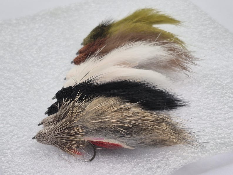 May include: A selection of fishing flies in shades of brown, black, white, and olive green. Each fly features a textured body and a small hook, designed for angling.