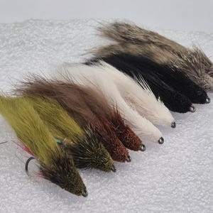 May include: A selection of handmade fishing flies in a range of colours, such as olive green, brown, white, and black. Each fly has a hook and is made with textured materials, probably to attract fish. The flies are displayed on a white surface.