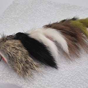 May include: A selection of fishing lures designed to resemble small creatures. The lures are made from fur in various colours, including brown, black, white, and olive green, each with a hook. They are arranged on a white surface.