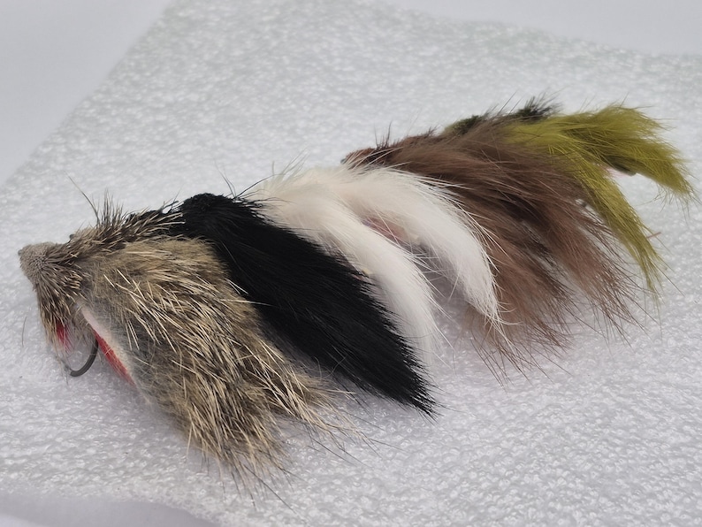 May include: A selection of fishing lures designed to resemble small creatures. The lures are made from fur in various colours, including brown, black, white, and olive green, each with a hook. They are arranged on a white surface.