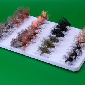 May include: A white foam fly-tying box containing 20 different fly patterns. The flies are arranged in rows and are a variety of colours, including brown, black, green, and orange.