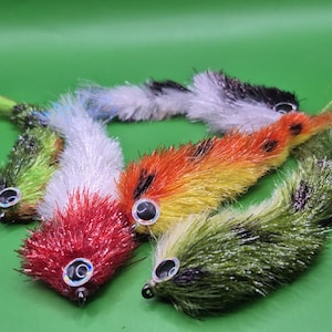 May include: A collection of colorful fishing lures with fluffy, textured bodies and large, reflective eyes. The lures are in shades of red, orange, yellow, green, white, and black, arranged against a green background. These are designed for fly fishing.