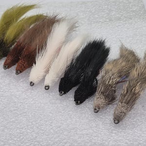 May include: A selection of fly fishing lures in shades of green, brown, white, black, and grey. Each lure features a textured body and a small metal eyelet at the front.
